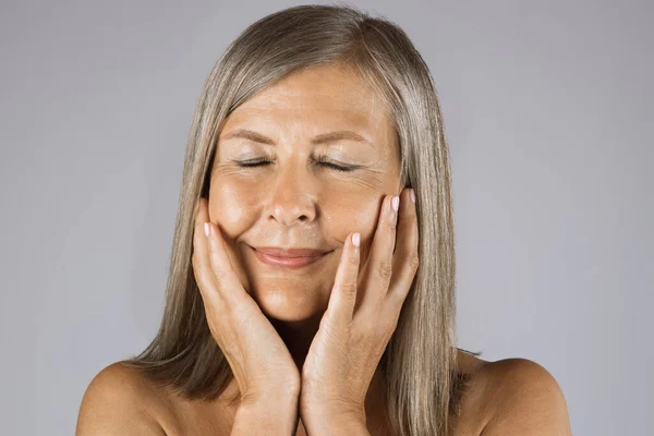 Aged woman keeping eyes closed and touching cheeks