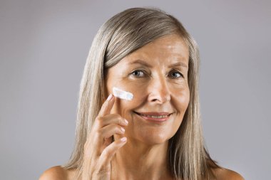 Yaşlı bir kadın parmaklarıyla yüzüne nemlendirici krem sürüyor.