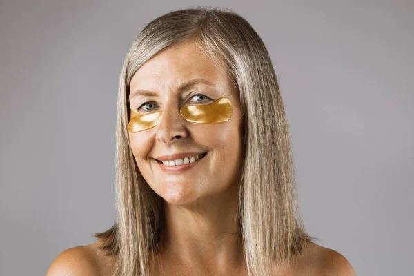 Portrait of aged woman using collagen eye patches in studio — Stock Photo, Image