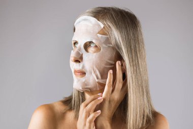 Yüzünde maskeyle gri arka planda poz veren yaşlı bir kadın.