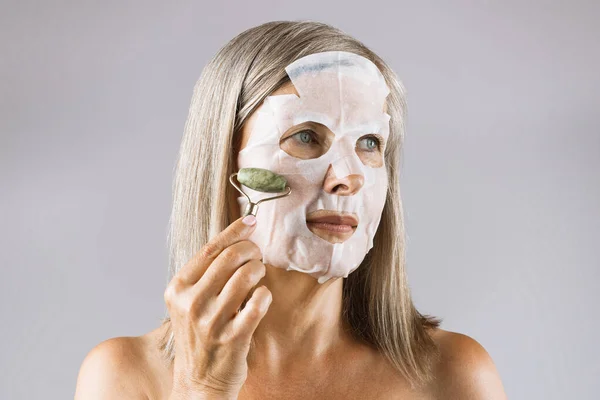 Starší žena v masce na obličeji dělá masáž s nefritovým válečkem — Stock fotografie