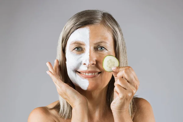 Aged woman with mask on face holding slice of cucumber — Stock Photo, Image