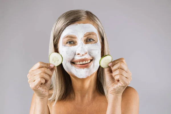 Pozytywna kobieta z maską na twarzy trzymająca plasterki ogórka — Zdjęcie stockowe
