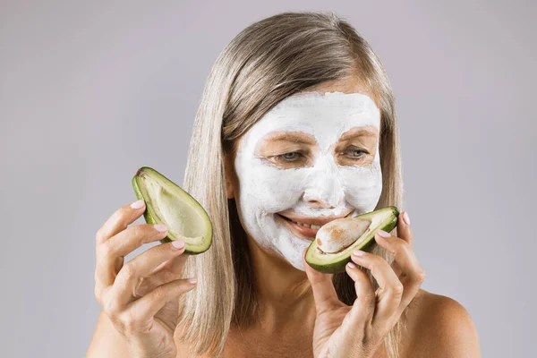 Stará žena s kosmetickou maskou na obličeji drží avokádo — Stock fotografie