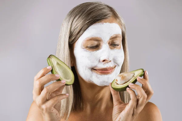 Wiek kobieta z maską kosmetyczną na twarzy gospodarstwa awokado — Zdjęcie stockowe
