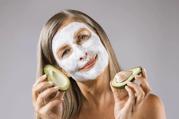 Женщина в маске для лица держит половинки авокадо в студии — стоковое фото