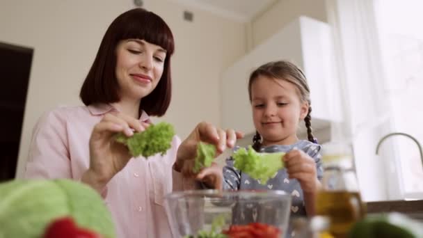 Tochter hilft ihrer Mama, frisch gewaschene frische Salatblätter in eine separate Glasschüssel zu reißen — Stockvideo