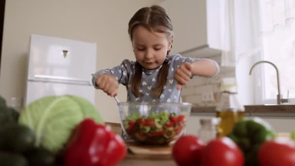 Mała dziewczynka spędza wolny czas mieszając świeże posiekane warzywa w szklanej misce przygotowując sałatkę. — Wideo stockowe