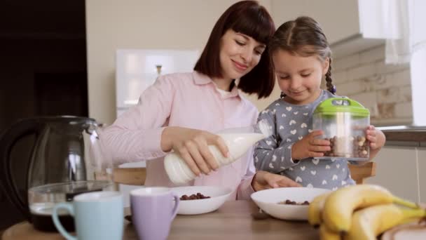 Νεαρή μητέρα ρίχνει γάλα σε κόρες φιόγκους από κορν φλέικς για πρωινό στο φως σύγχρονη κουζίνα. — Αρχείο Βίντεο