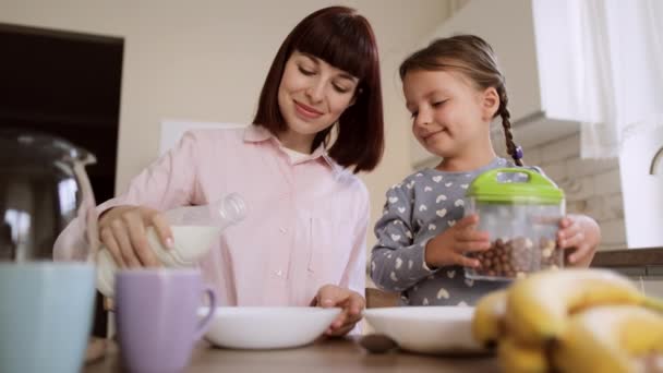 Ung mor häller mjölk i döttrar bågar majsflingor till frukost i ljust modernt kök. — Stockvideo