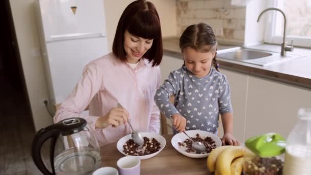 Fiatal anya és boldog kis aranyos lány ül a konyhaasztalnál, és élvezi a reggelit — Stock videók