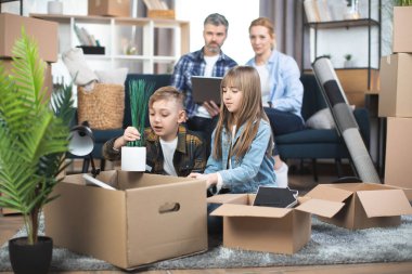 Çocuklar yeni dairede kutu açıp oynaşırken ebeveynler kanepede dizüstü bilgisayarlarıyla oturuyorlar.