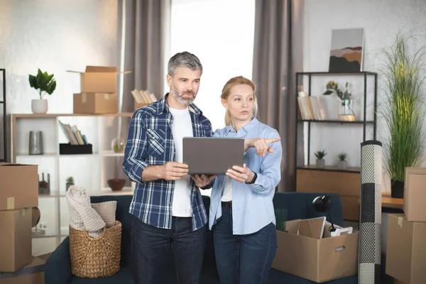 Hombre maduro y su hermosa esposa usando el ordenador portátil mientras se traslada a un nuevo piso — Foto de Stock