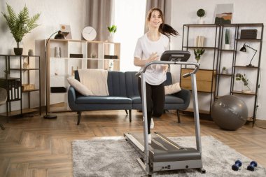 Siyah spor taytlı ve beyaz tişörtlü genç bir kadın evdeki koşu bandında koşuyor..