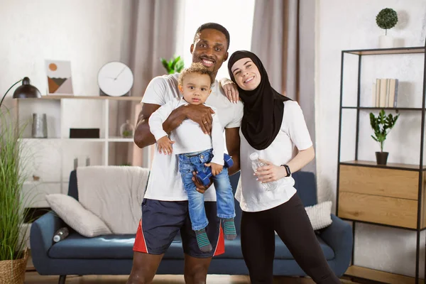 Homme africain tenant son fils dans ses mains et femme musulmane debout et souriant après l'entraînement domestique. — Photo