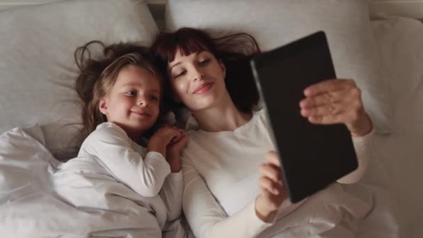 Madre y su hija tumbadas en la cama, divirtiéndose con la tableta digital. — Vídeos de Stock