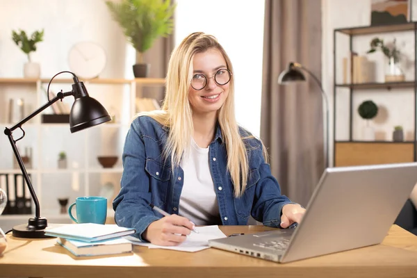 Usmívající se blondýnka psaní poznámek při používání notebooku, sedí za stolem doma — Stock fotografie
