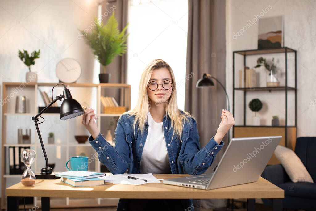 Female freelancer in eyeglasses meditating at table, feeling relaxed