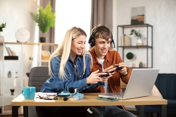 Genç mutlu çift evde bilgisayar oyunu oynuyor, dizüstü bilgisayarla masada oturuyor ve joystick kullanıyor. — Stok fotoğraf