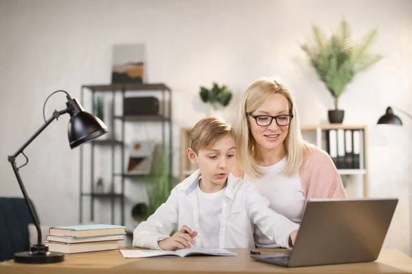 Молода мати в окулярах вказує на ноутбук, пояснюючи урок маленькому синові — стокове фото