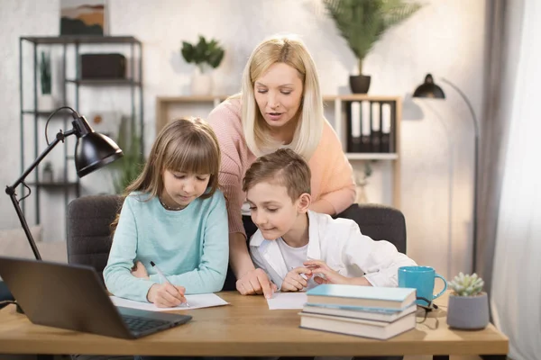 Mother teach young son and daughter homework, using digital laptop at home. — стокове фото