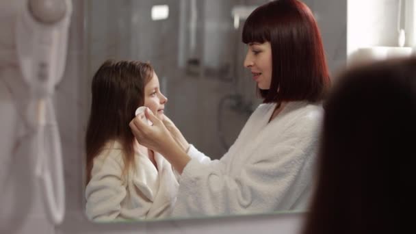 Jonge moeder in wit badjas reinigt met katoen pads gezicht van haar schattig mooi kind meisje. — Stockvideo