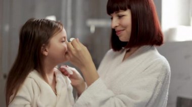 Beyaz bornozlu genç anne güzel kızının yüzünü pamuk pedlerle temizliyor..