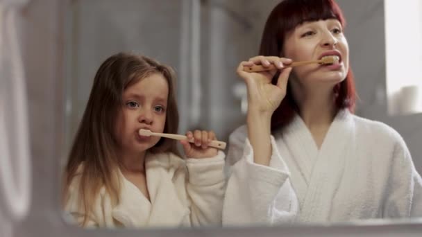 Mom and her cute girl in white bathrobe brushing teeth together standing near the mirror. — Wideo stockowe