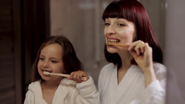 Mamá y su linda chica en albornoz blanco cepillarse los dientes juntos de pie cerca del espejo. — Vídeos de Stock