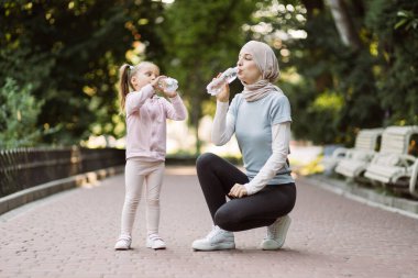 Arap anne kızına bakıyor ve spordan sonra dışarıda dinlenirken su içiyor.