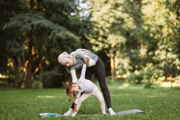Muslim woman in sportswear teaching her preschool cute daughter how to do side tilts in the park — Stock Photo, Image
