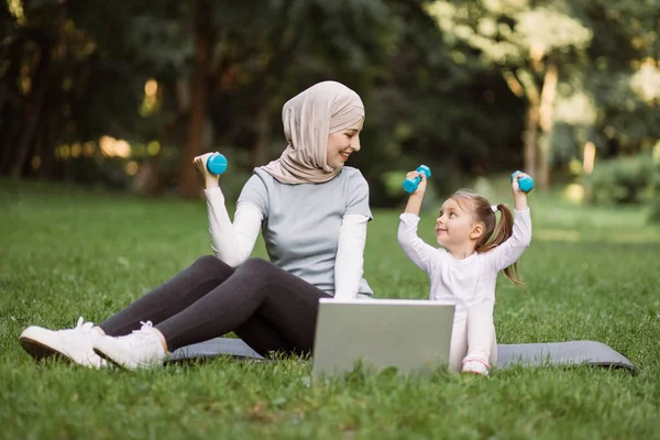 Mamá en hijab mostrando a su hijo cómo hacer ejercicio con pesas, viendo video en el portátil en el parque — Foto de Stock