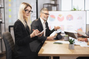 Modern ofis odasındaki masada dijital tablet ve kağıtlarla çalışan iki iş arkadaşı..