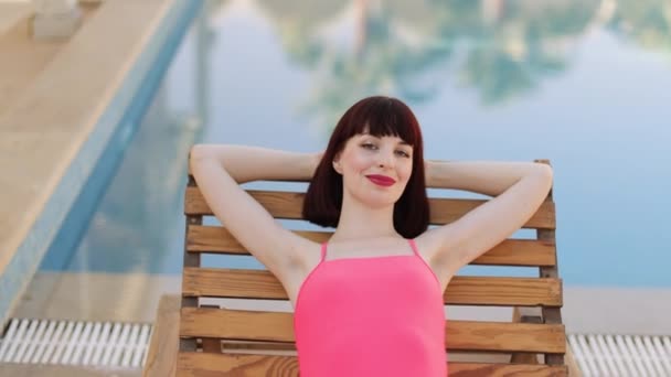 Pretty young woman lying on wooden deckchair near pool, put hands behind head — Stock Video