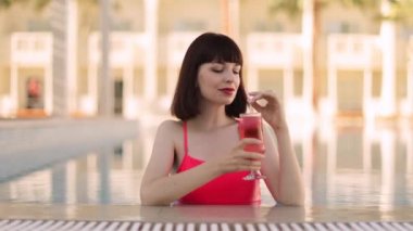 Pembe mayo giymiş güzel bir kadın havuzda yüzerken nefis bir kokteyl içiyor.