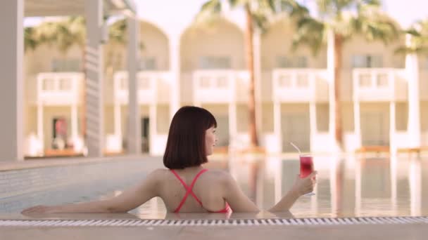 Pembe mayo giymiş güzel bir kadın havuzda yüzerken nefis bir kokteyl içiyor. — Stok video