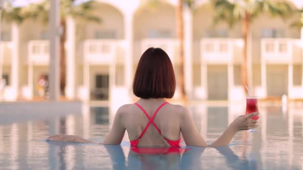 Mujer joven en sombrero elegante nadando en la piscina y disfrutando de un delicioso cóctel — Vídeo de stock