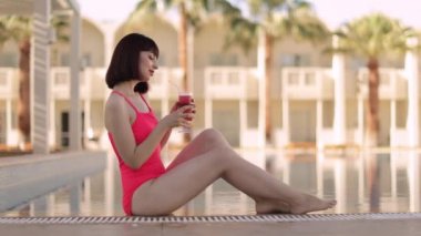 Pembe mayo giymiş bir kadın taze bir kokteylin tadını çıkarıyor, modern lüks deniz tatil beldesinde mavi bir havuzun kenarında oturuyor.
