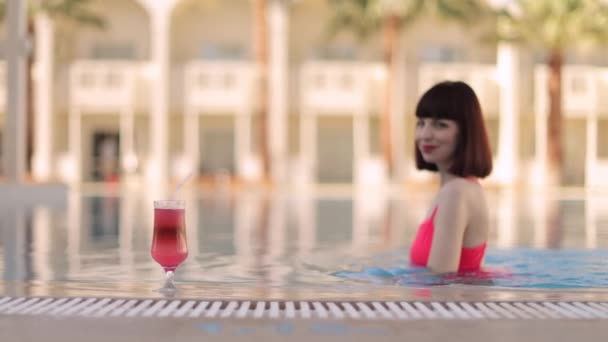 穿着鲜艳粉色泳衣的女人，在度假胜地的游泳池里喝着新鲜的五彩缤纷的鸡尾酒 — 图库视频影像