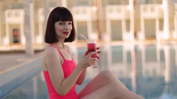 Жінка в елегантному рожевому купальнику, розслабляється біля басейну в сонячний день, з освіжаючим фруктовим коктейлем . — стокове відео