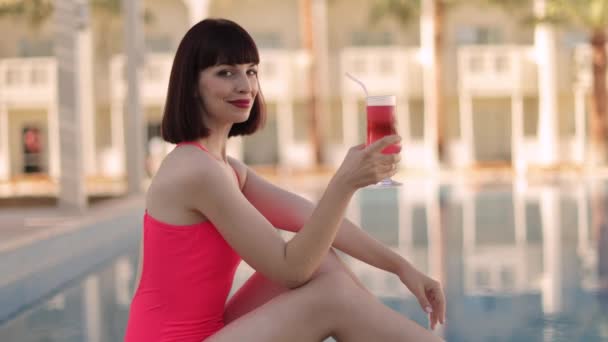 Donna in costume da bagno rosa, seduta davanti alla piscina del resort di lusso, che beve cocktail — Video Stock