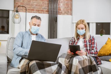Kanepede dizüstü bilgisayar ve tablet kullanan maskeli olgun bir çift.