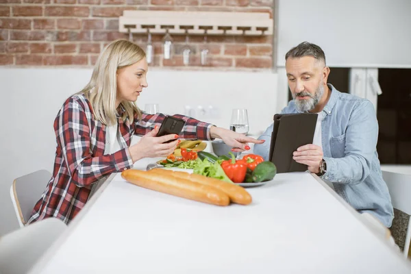 Mature couple sitting at kitchen table with gadgets in hands — Stock Fotó