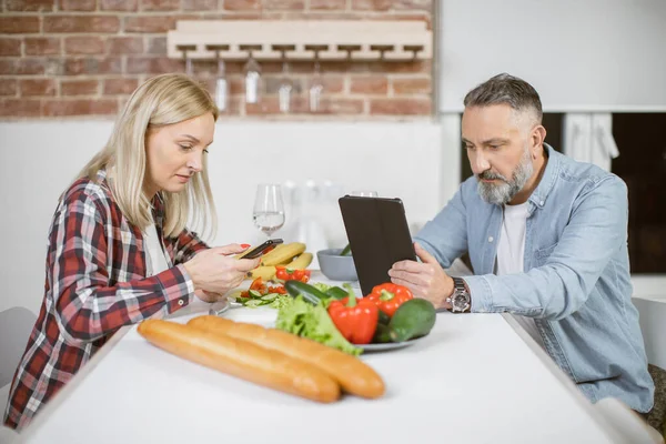 Mature couple sitting at kitchen table with gadgets in hands — Stock Fotó