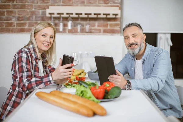 Portrait of couple sitting on kitchen with gadgets in hands — стоковое фото