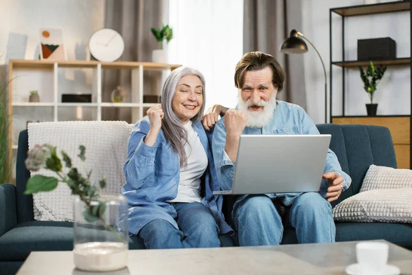 Χαρούμενα ηλικιωμένα ζευγάρια gesturing για εκφράζοντας την ευτυχία, ενώ κάθεται στον καναπέ με φορητό υπολογιστή. — Φωτογραφία Αρχείου