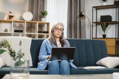 Evdeki oturma odasında video sohbet için kulaklık ve dizüstü bilgisayar kullanan güzel bir kadın..