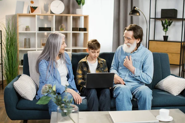 Εγγονός διδάσκει τους παππούδες του να χρησιμοποιούν το φορητό υπολογιστή κάθεται στον καναπέ στο σαλόνι. — Φωτογραφία Αρχείου