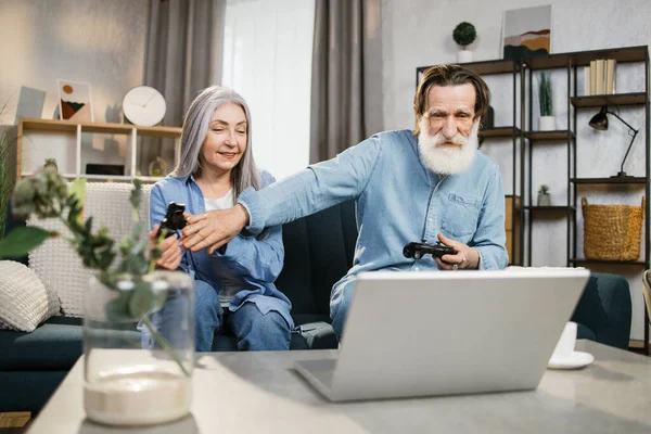 Ευτυχισμένη ώριμη οικογένεια που παίζει βιντεοπαιχνίδια στο σπίτι, κάθεται στον καναπέ με φορητό υπολογιστή και χρησιμοποιεί joysticks. — Φωτογραφία Αρχείου