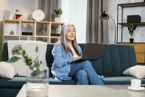 Neşeli olgun beyaz kadın koltukta oturuyor ve kablosuz bilgisayarla çalışıyor..